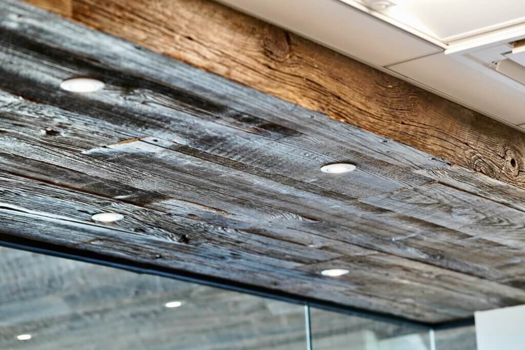 A low-angle, close-up photo shows a ceiling and wall area covered with reclaimed antique wood. A large, rough-hewn brown timber beam runs along the top edge of the frame. Below it, the ceiling is clad in weathered gray reclaimed wood planks, into which several small, white recessed lights are installed. The wood has visible texture, knots, and nail holes.